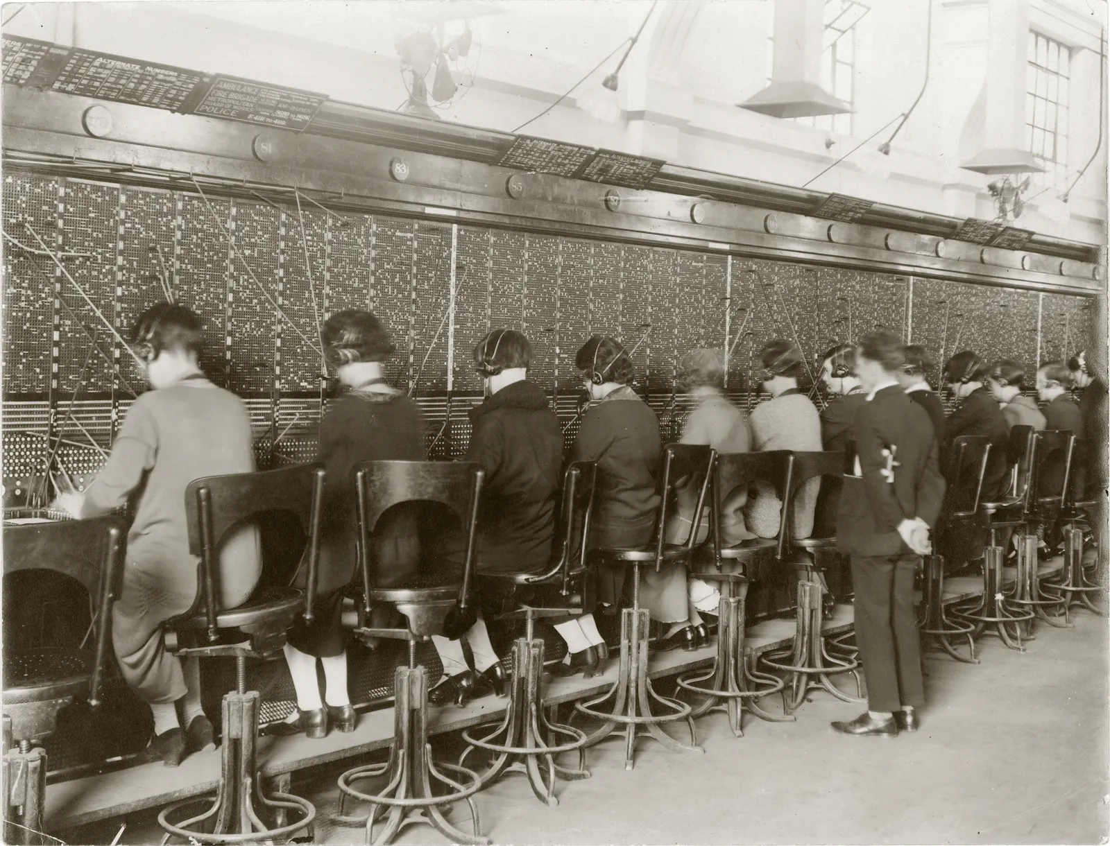 A switchboard running the length of a room, its face a dense field of jacks and lamps with cords looping up from the shelf; fourteen telephonists in headsets sit in a row on high swivel stools with their backs to the camera, and a young man in a dark suit stands among them, hands behind his back. Numbered brass discs run along the top of the board, and a sign above it lists the numbers for the ambulance, the fire brigade, the Board of Works and the police.