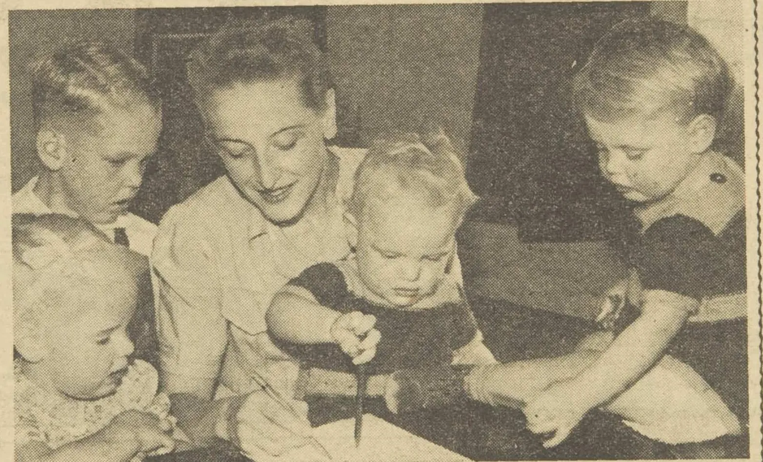 A newspaper photograph of a smiling woman at a table with four small children, one of them, a toddler, holding a pen to a sheet of paper.