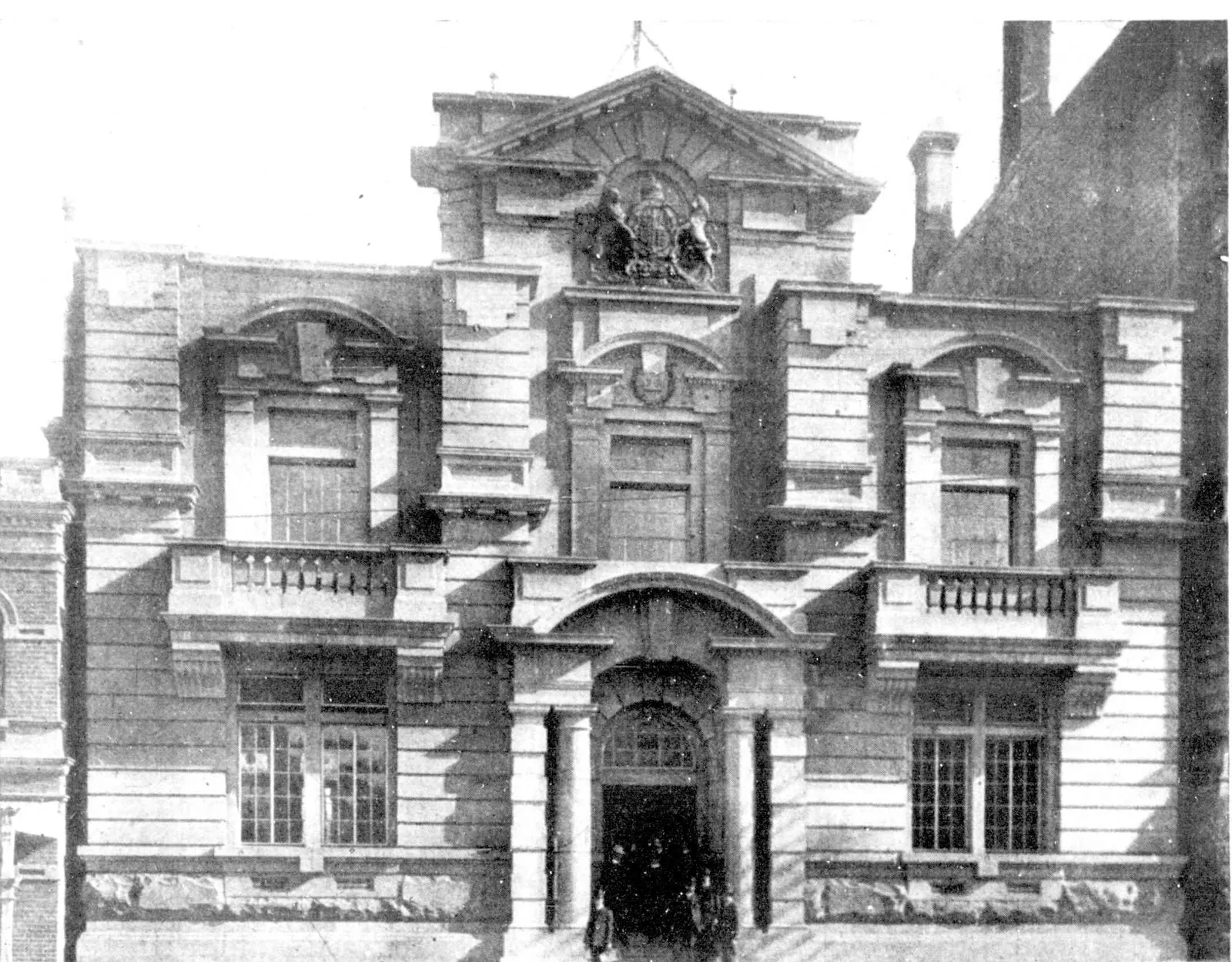 A newspaper photograph of a two-storey stone building with a carved coat of arms on its pediment, arched upper windows with balconies, and a columned doorway on the street.