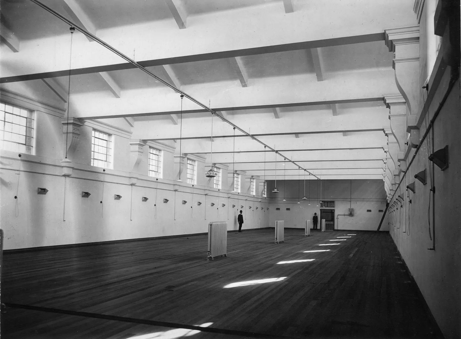 A long empty hall with a beamed concrete ceiling, tall windows down one wall, bare floorboards striped with sunlight and cable outlets along the walls; two men in dark suits stand far down the room.